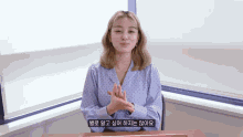 a woman in a blue polka dot shirt is sitting at a desk with her hands folded in front of a window