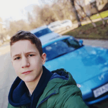 a young man in a green jacket is taking a selfie in front of a blue car