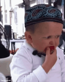 a little boy wearing a hat and a bow tie is eating a red apple .