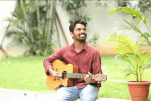 a man in a red shirt is holding a guitar in front of a potted plant