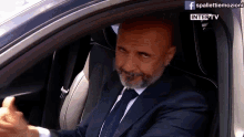 a man in a suit and tie is sitting in the driver 's seat of a car with a facebook logo behind him