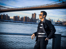 a man wearing sunglasses and a striped shirt stands in front of a bridge over a body of water