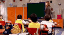 a group of children are sitting at desks in a classroom with a teacher standing in front of a blackboard .