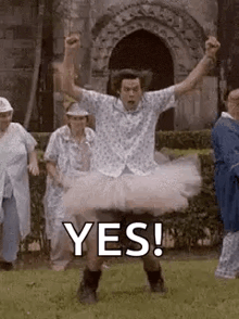 a man in a tutu is standing in the grass with his arms in the air and a group of people behind him .