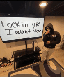 a man in a hoodie stands in front of a tv screen that says " lock in yk i want you "