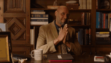 a man sits at a desk in front of a bookshelf with a mug that says ' i 'm sorry '