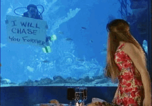 a woman in a red dress stands in front of an aquarium holding a sign that says i will chase you forever