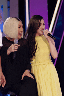 a woman in a yellow dress is singing into a microphone next to another woman in a black dress