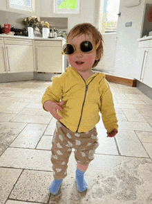 a little girl wearing sunglasses and a yellow jacket is standing on a tiled floor