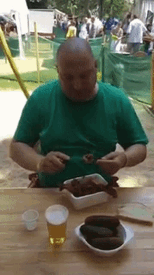 a man in a green shirt is sitting at a table with a bowl of pickles and a glass of beer