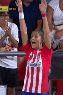 a woman in a red and white striped shirt is raising her arms in the air while watching a soccer game .