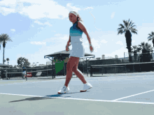 a woman walking on a tennis court with palm trees in the background and a sign that says ' wilson ' on it