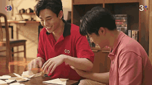 two young men are sitting at a table playing with paper and one of them is wearing a red shirt with the letters go on it