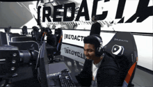 a man wearing headphones sits at a desk in front of a sign that says ' redact ' on it