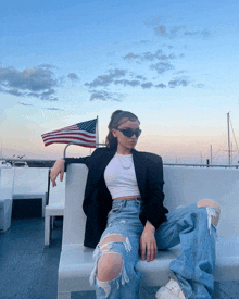 a woman wearing ripped jeans and sunglasses sits on a bench with an american flag in the background