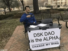 a man is sitting at a table with a sign that says dsk dao is the alpha change my mind