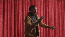 a man in a brown jacket is standing in front of a red curtain looking at his phone