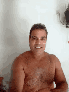 a shirtless man smiles for the camera while sitting in front of a white wall