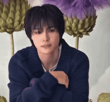 a young man in a blue sweater is sitting in front of purple flowers