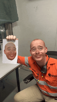 a man in an orange shirt holds up a piece of paper with a picture of his face on it