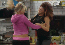 two women are fighting in a kitchen and one of them is wearing a pink hoodie