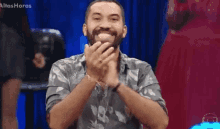 a man with a beard is smiling and clapping his hands while sitting in front of a blue curtain .