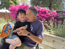 a man in a purple shirt is holding a young boy in front of pink flowers