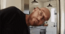 a bald man with glasses is standing in a kitchen with his head turned to the side .