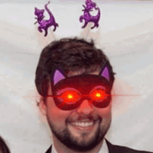 a man with a beard is wearing a cat mask with red eyes and a purple cat headband .