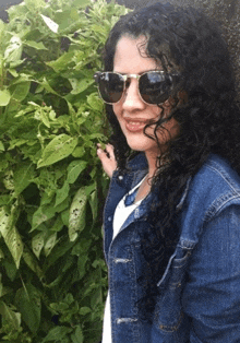 a woman wearing sunglasses and a denim jacket is standing in front of a bush