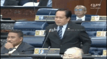 a man in a suit and tie is giving a speech in front of a microphone in a parliament chamber