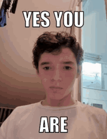 a young boy with curly hair is wearing a white t-shirt and a meme .