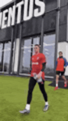 a man in a red shirt is walking on a soccer field in front of a building that says juventus