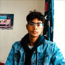 a young man wearing glasses and a denim jacket is sitting in a room .