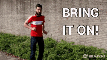 a man in a red adidas shirt is standing in front of a brick wall with the words bring it on written on it