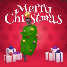 a man dressed as a christmas tree is standing in front of gifts and the words merry christmas