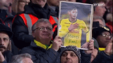 a man in a crowd holds up a picture of a man in a yellow jersey that says merci emi