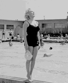 a woman in a black bathing suit is standing on the edge of a swimming pool holding a hat .