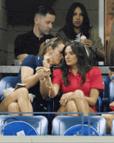 a woman in a red polo shirt sits in a stadium talking to another woman