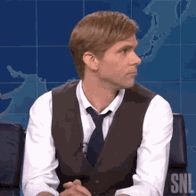 a man wearing a vest and tie is sitting in front of a snl logo