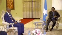 two men sit in chairs in front of a blue and white flag and a table with bottles on it