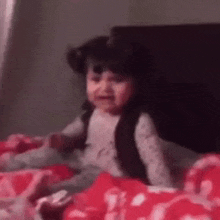 a little girl is crying while sitting on a bed with a red blanket .