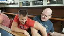 two men are sitting at a table with one wearing a red shirt