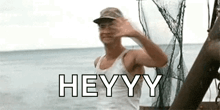 a man in a white tank top and hat is waving at the camera while standing on a boat in the ocean .