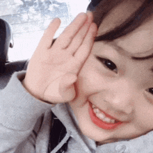 a little girl is smiling and waving at the camera with her hand on her face .