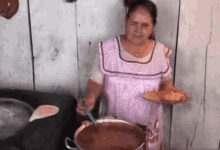 a woman in a pink apron is holding a plate of food in front of a large pot of soup .