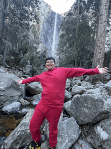 a man in a red hoodie stands in front of a waterfall in the woods