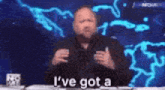 a man is sitting at a table talking into a microphone and saying `` i have got a '' .