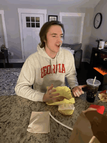 a man wearing a georgia sweatshirt sits at a table eating a hamburger