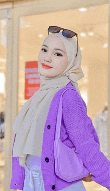 a woman wearing a purple cardigan and a white hijab is standing in front of a store .